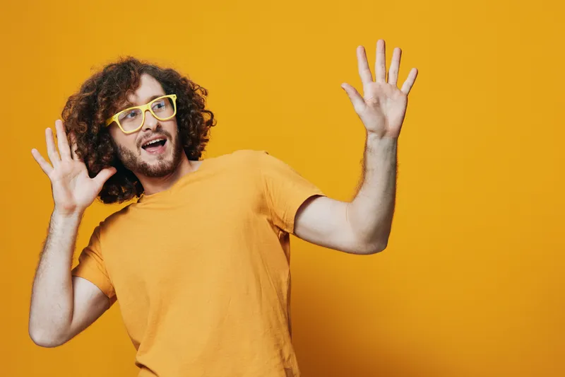 Cheerful young man with curly hair in yellow glasses expressing joy against a bright yellow background, perfect for creative projects and vibrant marketing