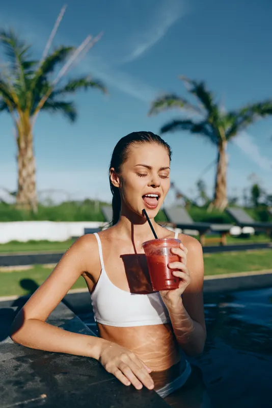 Young woman in a white bikini top relaxes in a pool, sipping a bright smoothie through a straw, palm trees and sunshine create a vibrant lifestyle moment by the water