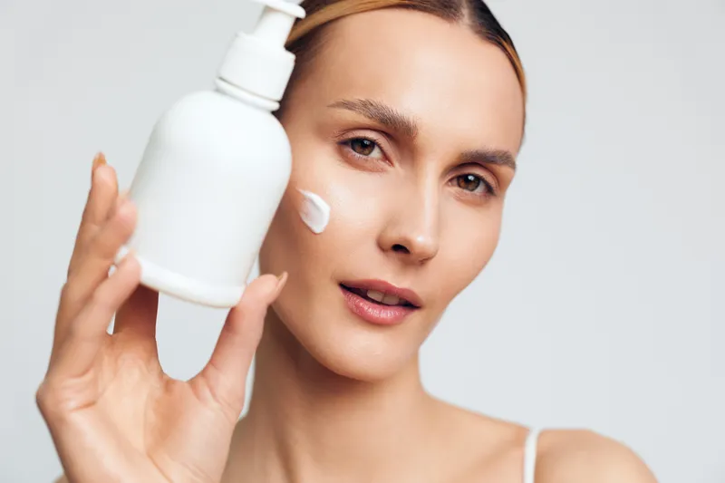 Young woman applying cream on face holding white bottle close up skincare concept natural beauty clean skin soft light minimal background