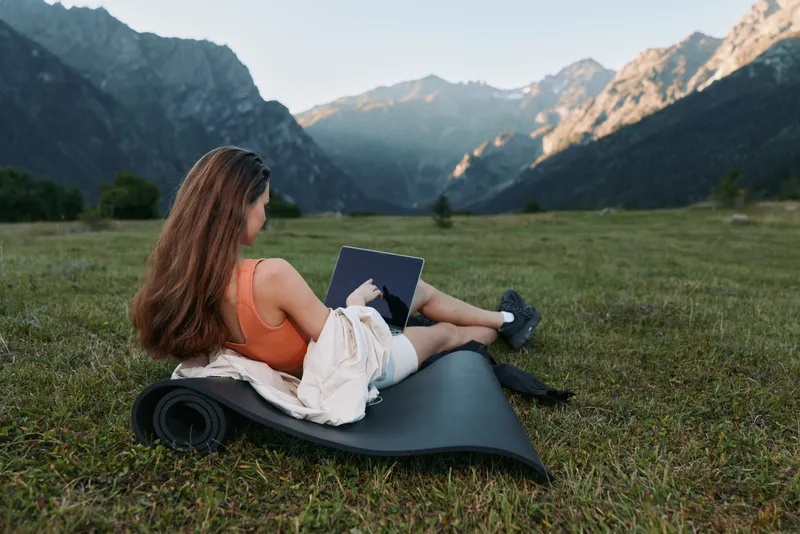 A woman sits on a rolled yoga mat in a grassy meadow beneath towering mountains. She works on a laptop in casual attire, looking focused yet relaxed in a serene outdoor setting.
