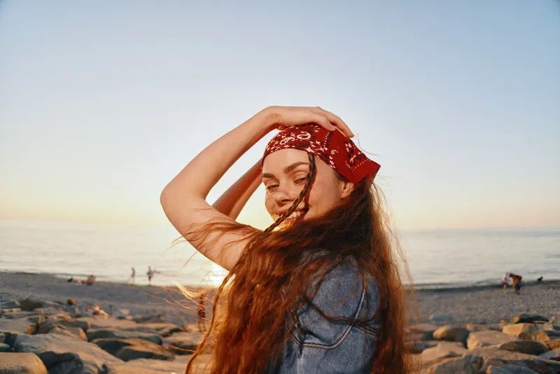 Lifestyle woman with modern boho-western denim street style poses joyfully by the beach at sunset, embracing natural film color and carefree summer vibes.