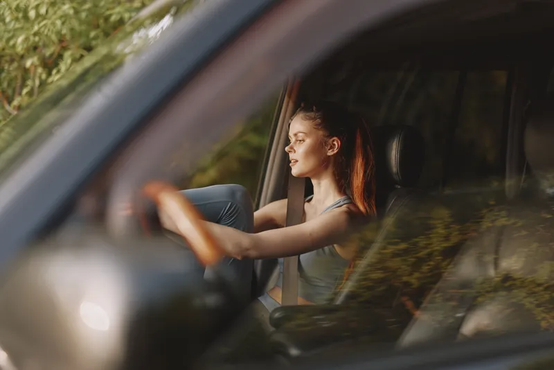 Young woman driving a car with a relaxed expression, showing confidence and joy of the journey, surrounded by nature in a warm color palette