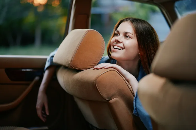 Happy young woman enjoying a car ride with a bright smile, wearing casual clothing, captured in a warm and inviting setting at sunset, reflecting joy and freedom.