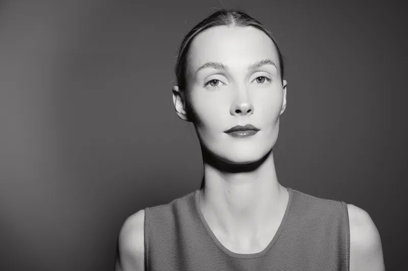 Portrait of a fair skinned Caucasian woman with slicked back hair wearing a sleeveless top against dark background. Minimalist beauty concept.
