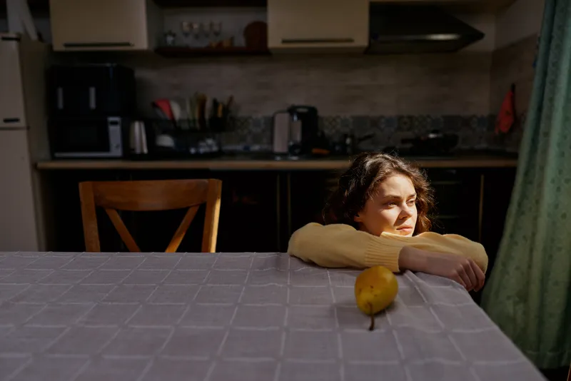 A pensive woman rests her arms on a kitchen table, gazing into the distance. A lemon sits at the edge, suggesting quiet domestic reflection and solitude.