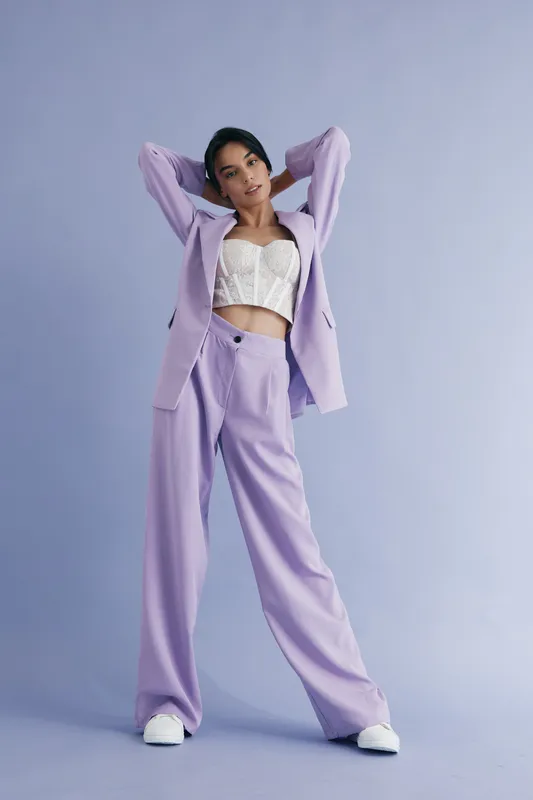 Stylish woman in lavender suit and white lace top posing confidently against a pastel purple background, showcasing modern fashion and elegant attitude.