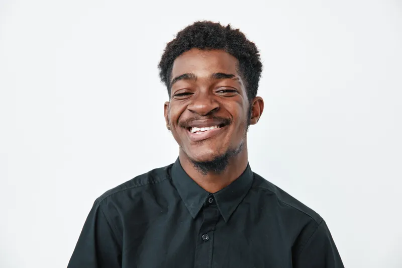 Young black man with dark skin smiling and wearing black shirt on white background. Portrait of happy African ethnicity male with short curly hair and closed eyes.