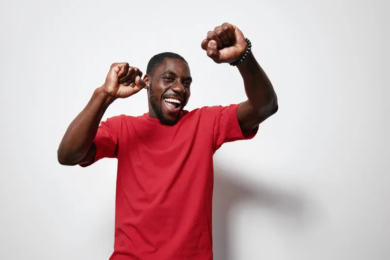 African American man with dark skin wearing red t shirt dancing and smiling with raised fists on white background for celebration and happiness concept.