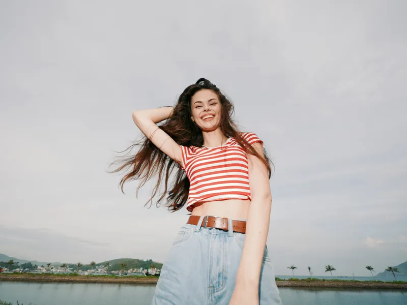 Bright portrait of a smiling young woman in a striped crop top and denim jeans, posing confidently with wind in her hair against a coastal sky.