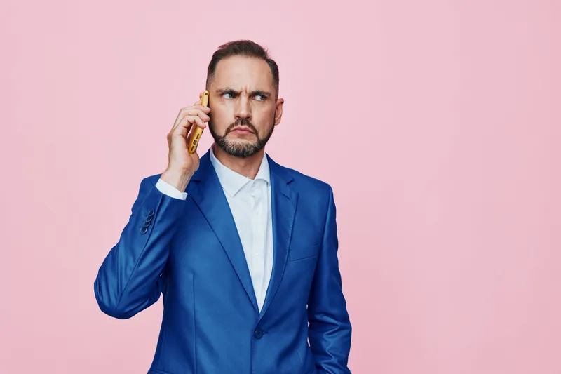 A professional man in a blue suit on a pink backdrop, speaking on a mobile phone. The isolated colored setting highlights corporate mood and a focused expression.