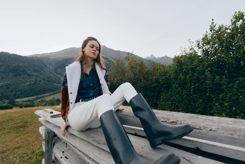woman in nature with mountains and boots posing in outdoor fashion, sitting on wooden bench in countryside meadow, casual denim jacket and white trousers for autumn style and relaxation