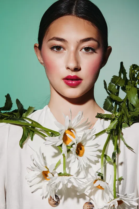 Young woman with vibrant pink lipstick and flawless skin poses with white daisies hanging from greenery. Bright studio lighting highlights fresh makeup and creative fashion details. Beauty look.