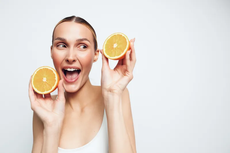 Young woman with orange halves smiles playfully in a bright studio, the citrusy colors reflecting her energetic mood, relaxed posture, and fresh sunlit vibe.