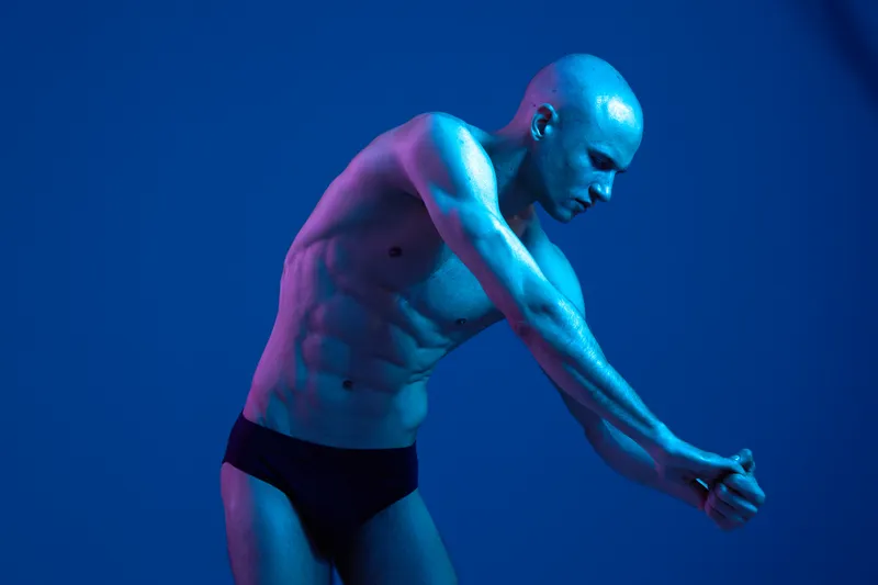 Athletic man posing in dynamic posture, showcasing strength and flexibility against a vibrant blue background. The image vividly captures physical prowess and artistic expression. ###