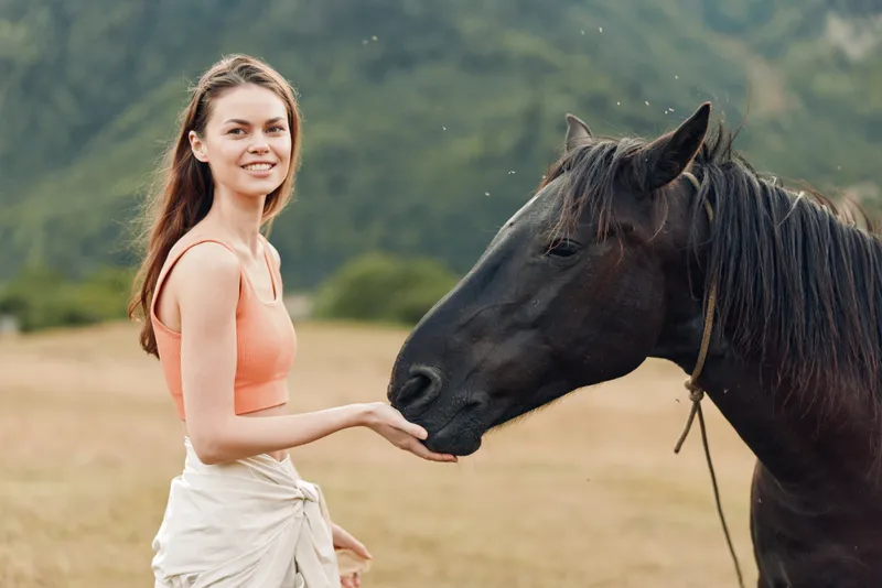 A young woman in a coral top gently touches a dark horse in a rural field, smiling softly as the animal nuzzles her hand, creating a warm, natural bond between human and horse.