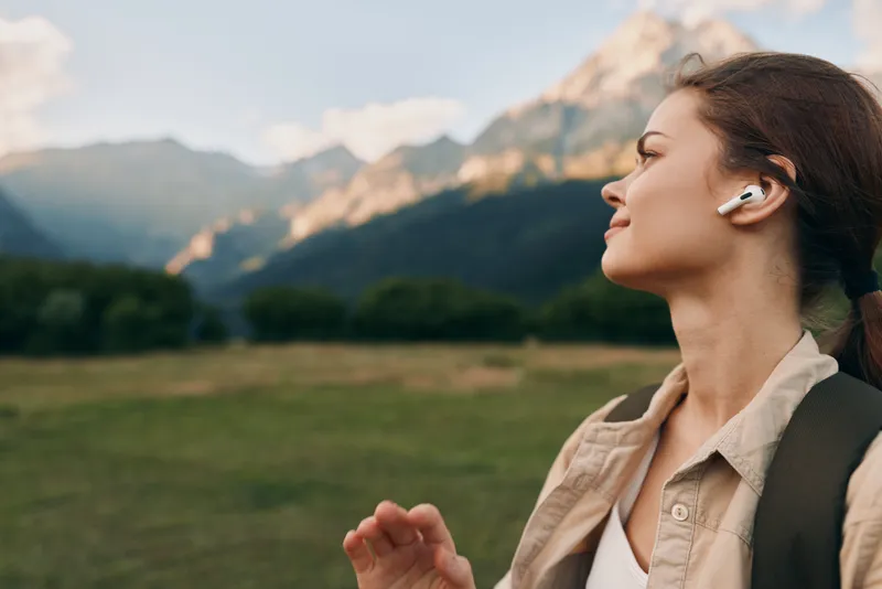 A young woman in a beige jacket with a backpack runs in profile, wearing wireless earbuds against a backdrop of mountains and open fields with soft sunlight.