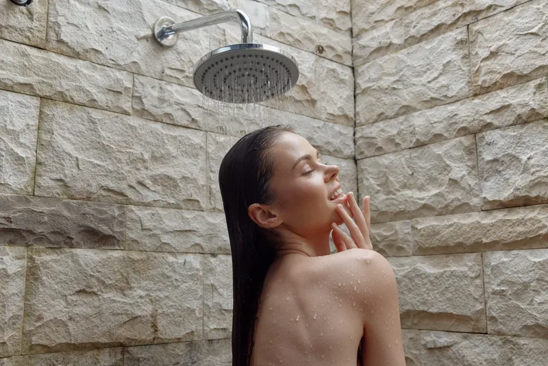 young woman enjoying outdoor shower with stone wall background, wet hair, relaxed and peaceful expression, natural skin tones, morning light