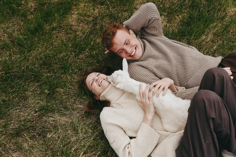 Joyful couple lying on grass with a goat kid, enjoying an outdoor lifestyle, bonding and happiness in a relaxed moment, soft light, cozy sweaters, close connection, nature and playful calm.