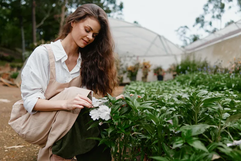 Young woman caring for flowers in a greenhouse, wearing casual clothing, surrounded by greenery, showcasing the beauty of nature and sustainable gardening