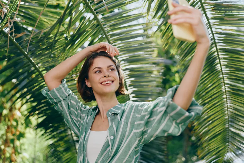 Young woman in a striped green shirt takes a selfie under palm fronds, smiling as she captures a sunny outdoor moment.