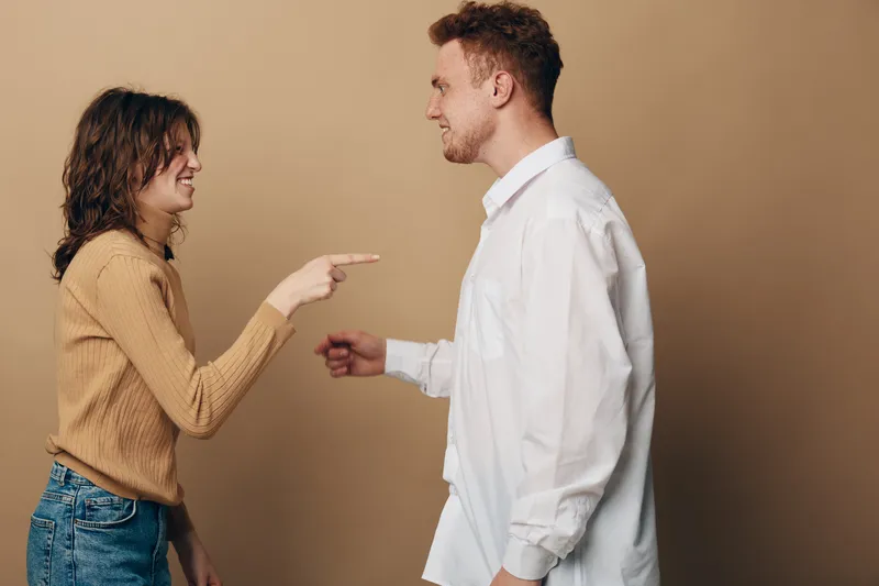 Young woman in brown sweater points finger at smiling man in white shirt. Casual interaction between friends or couple on beige background. Concept of communication and emotions.