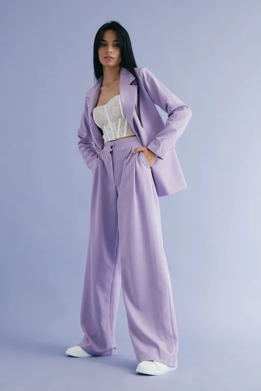 Young woman wearing lilac pantsuit and white lace top posing confidently against a pastel purple background, showcasing modern fashion and casual elegance.