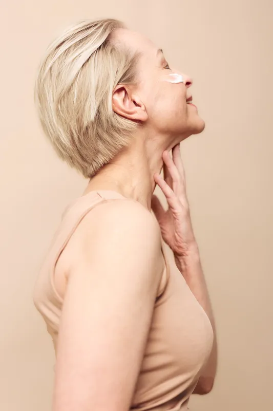 Smiling mature woman with short blonde hair applies white facial cream on cheek, touching neck gently. Beauty and skincare concept with natural skin care, wellness, and self-care routine.