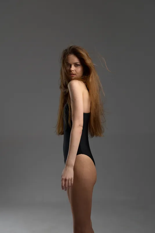 slender woman with long hair posing in black bodysuit against gray background, minimalistic fashion portrait