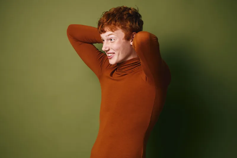 Young redhead woman with short hair wearing brown turtleneck smiling and looking to side against green background with hands behind head and relaxed pose