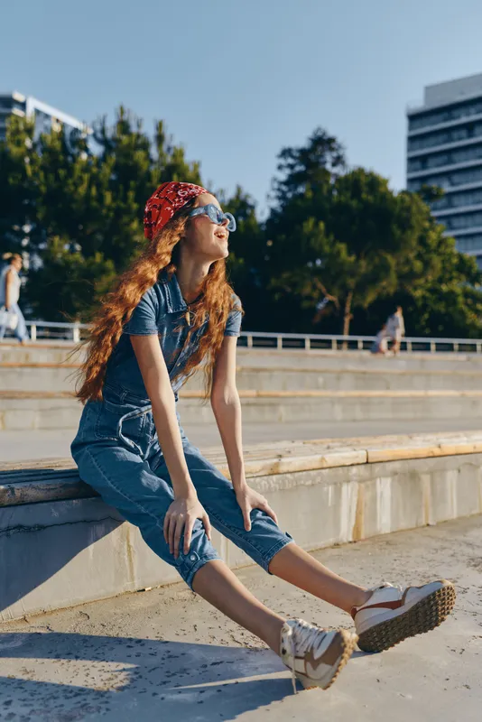 Lifestyle woman in modern boho-western denim street style sitting outdoors, wearing red bandana and sunglasses with film color effect on a sunny day.