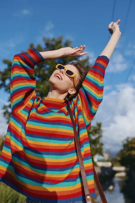 Woman rainbow sweater sunglasses raises arms outdoors, happy portrait of a stylish woman smiling under blue sky with bag strap, colorful knit fashion, joyful carefree mood and sunshine.