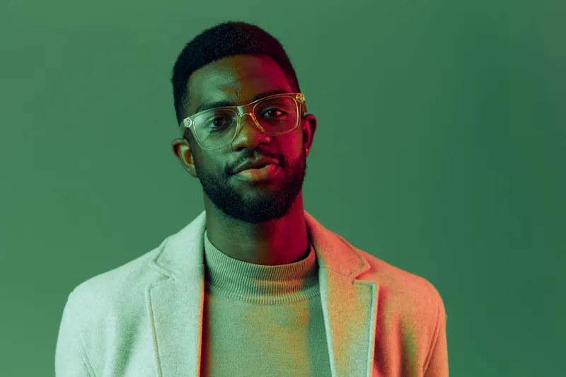 Stylish young African American man with glasses in a soft pastel outfit, showcasing confidence against a vibrant green background, perfect for fashion or lifestyle themes