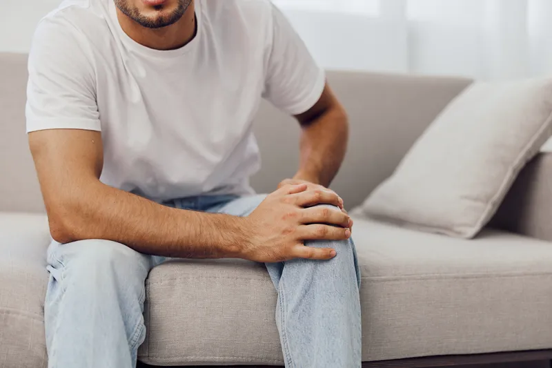 Casual man sitting on a couch, holding his knee in discomfort. The modern interior emphasizes relaxation while showcasing the significance of self-care in daily life.