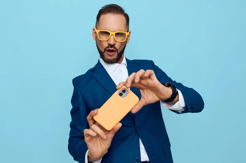 A professional businessman on a solid blue background captures a playful moment with a bright orange phone, expressing surprise, energy, confidence, and modern style.