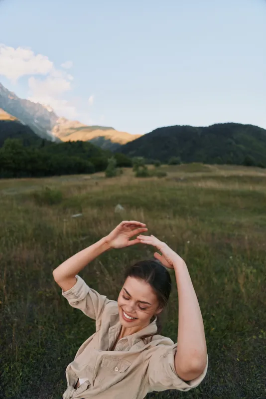 A happy woman in casual beige clothing enjoys nature with raised arms in a green meadow near forested hills under a clear sky. Lifestyle and outdoor concept.