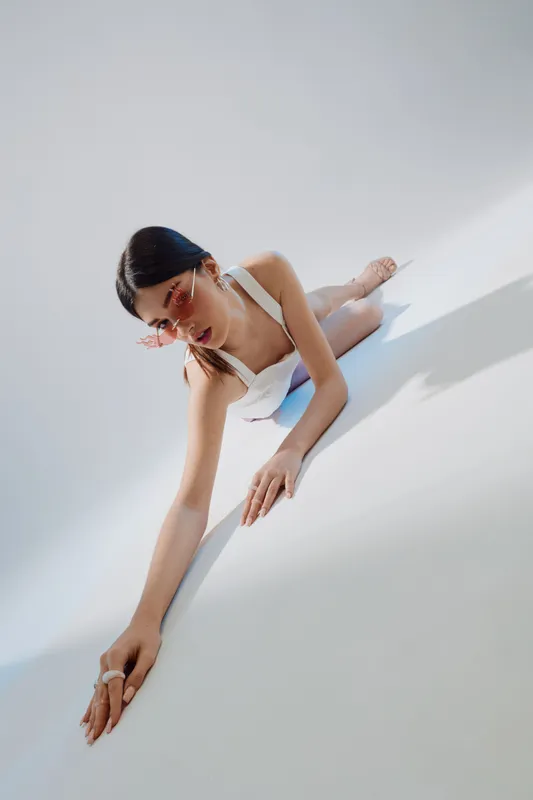 Young woman in white fashion outfit and pink sunglasses poses on white floor. Her long dark hair is styled back. Concept of beauty, fashion, and modern style photography.