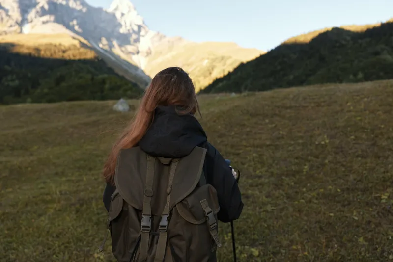 A lone hiker stands in a vast alpine meadow, facing distant rugged peaks. The subject wears a sturdy backpack and outdoor apparel, embodying exploration and outdoor adventure.