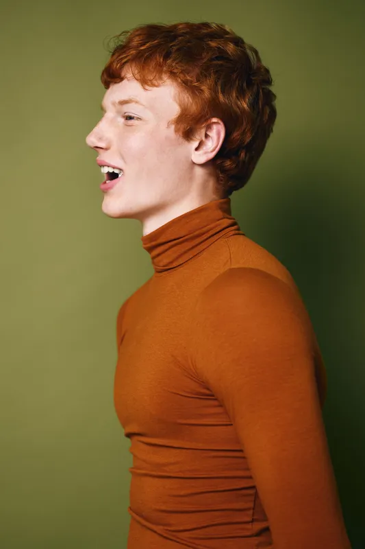 Young man with red hair wearing brown turtleneck shirt standing in profile view on green background with open mouth as if speaking or singing.
