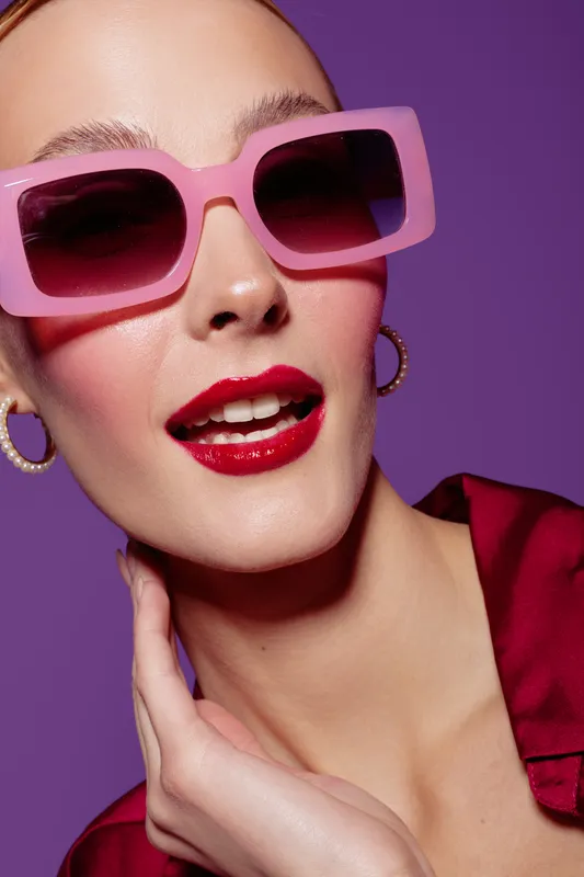 Close-up of fashionable woman wearing pink sunglasses and red lipstick posing against purple background. Concept of style, beauty, makeup, and confident attitude in studio lighting.