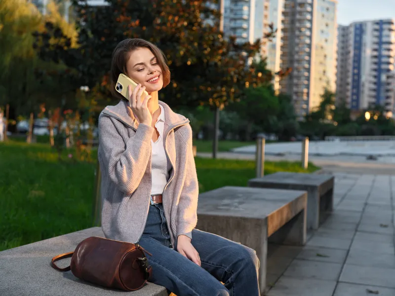 A cheerful young woman sits on a bench in a modern urban park, talking on a smartphone. She smiles warmly, enjoying a light moment outdoors, surrounded by trees and calm city ambiance.