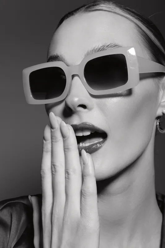 Close-up of a fair skinned Caucasian woman wearing oversized sunglasses and glossy lipstick with hand near mouth. Fashion, beauty, and style concept with studio lighting.
