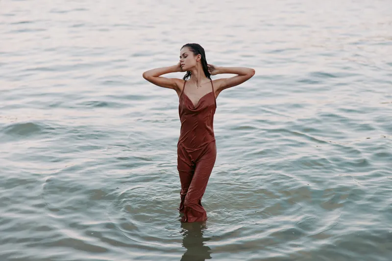 A woman stands waist deep in calm water, wearing a flowing red dress. Soft light highlights the fabric, creating a graceful silhouette and a tranquil, contemplative mood.