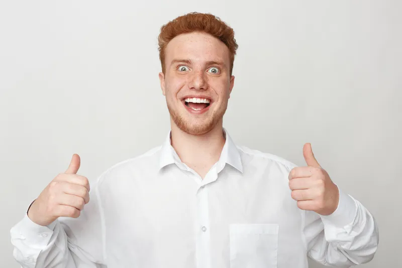 Happy young man with red hair and beard wearing white shirt shows thumbs up gesture with both hands. Positive facial expression on light grey background. Concept of approval and success.