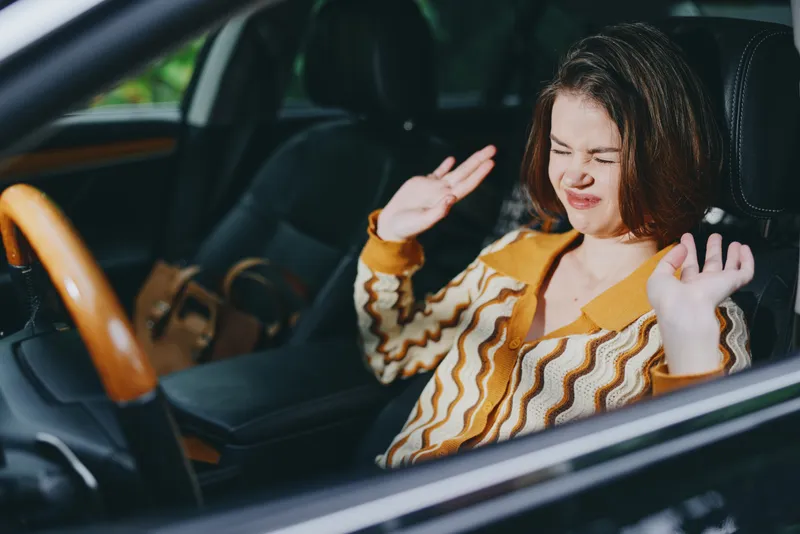 Young woman sits in a car, smiling and dancing with hands raised, enjoying music and a carefree moment behind the wheel.