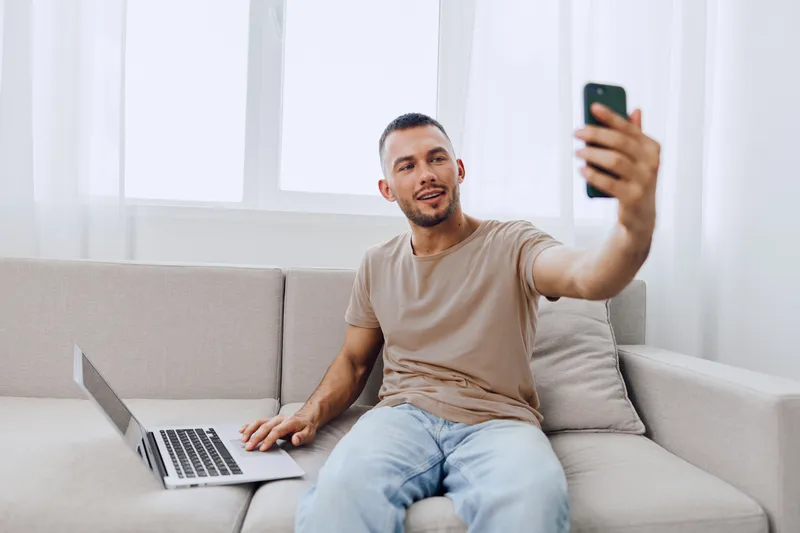 Motivated man taking selfie while video calling, showcasing modern technology and lifestyle trends, with a casual home environment and bright ambiance.