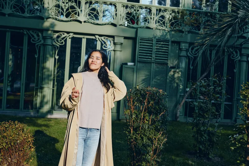 Woman in a long beige coat and jeans stands outdoors in sunlight, smiling, holding a drink, casual fashion scene in a residential garden setting.