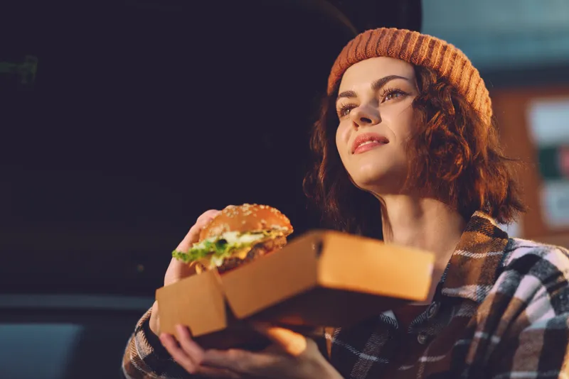 Woman holding burger in takeout box, wearing beanie in urban portrait with golden hour glow, authenticity and candid lifestyle vibe, mindful living and emotional storytelling.