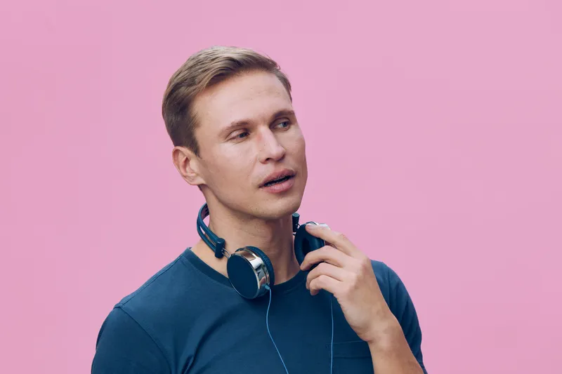 Young Caucasian man with short light brown hair wearing a blue sweater and headphones around his neck looks thoughtfully aside against a plain pink background, casual portrait, people lifestyle concep