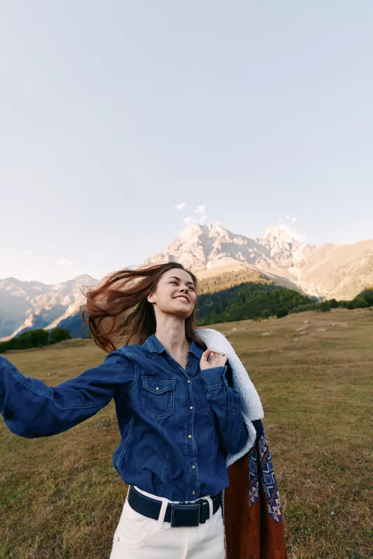 Woman smiling in denim shirt on mountain meadow outdoors, enjoying nature and breeze. Portrait of happy traveler with jacket and open alpine landscape in soft sunlight.
