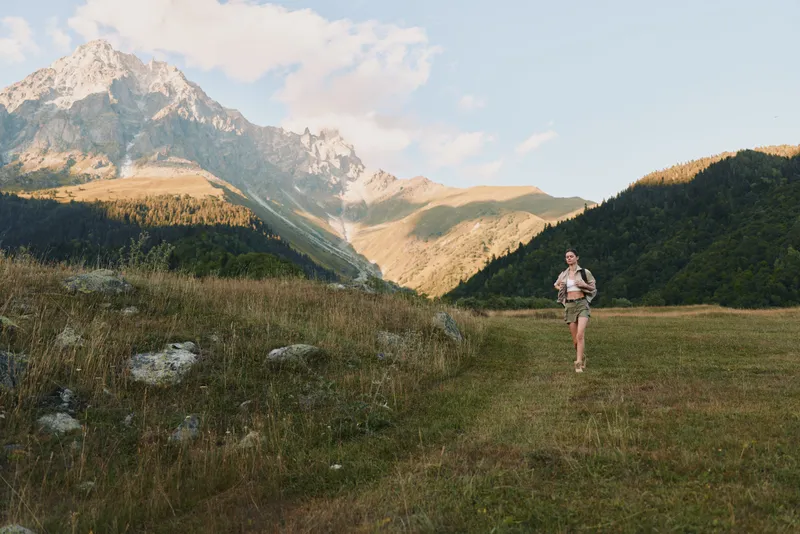 A woman in casual clothing runs through a grassy mountain meadow during daytime. The scene shows a clear sky and rocky peaks in the background, representing outdoor lifestyle.